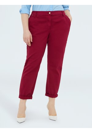 Persoon die opgerolde bordeauxrode broek, lichtblauw overhemd met knopen en witte schoenen met spits toelopende neus draagt, tegen een effen achtergrond.