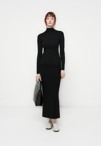 Svart ribbet turtleneck topp kombinert med et langt svart skjørt. Modellen holder en teksturert svart veske og har på seg lyse slip-on sko.