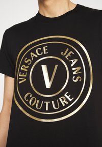 Schwarzes Baumwoll-T-Shirt mit einem großen goldenen Metallic-Logo mit "VERSACE JEANS COUTURE" und einem markanten "V" in der Mitte.