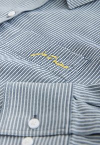 Pruhovaná modro-biela bavlnená košeľa s golierom na gombíky, žltou výšivkou a malými bielymi gombíkmi na légach.