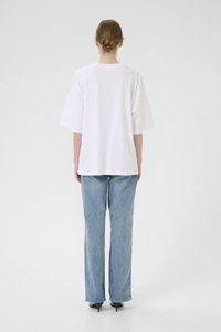 Tricou alb supradimensionat cu mâneci scurte, asortat cu blugi evazați de un albastru deschis. Fundalul este neutru și discret.