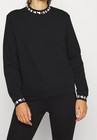 Sudadera negra con escote circular que presenta un detalle de marca en blanco. Mangas largas con puños acanalados y un ajuste holgado. Textura suave del tejido.