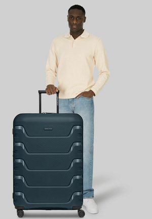 Hombre con camisa beige de manga larga y vaqueros junto a una gran maleta rígida azul oscuro con asa extendida sobre fondo gris.