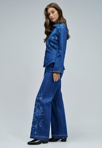 Veste en denim et jeans évasés en bleu profond, avec un motif floral subtil sur la jambe gauche du pantalon et des bottes à talons noires.