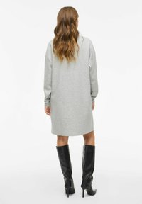 Vrouw met golvend bruin haar in een losse grijze jurk met lange mouwen en zwarte kniehoge hakken, staand met haar rug naar de camera tegen een witte achtergrond.