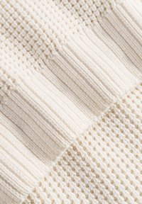 Primo piano di un tessuto lavorato a maglia di colore crema che mostra una varietà di texture, tra cui lavorazioni a coste e a nido d'ape, disposte in sezioni diagonali.