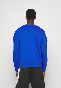 Personne debout de dos portant un sweat-shirt à manches longues bleu vif et un short noir avec des panneaux latéraux blancs sur un fond uni.