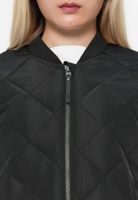 Quilted black bomber jacket with a front zipper, elasticated cuffs, and a smooth, shiny fabric texture. Features a round neckline.