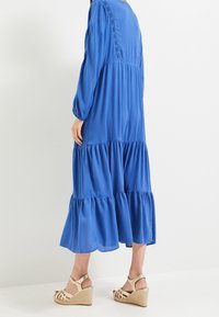 Dlouhé modré šaty vyrobené z lehké látky s vrstvenými volány, dlouhými rukávy a texturovaným zadním dílem. Doplňují je béžové platformové sandály.