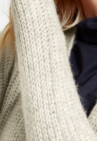 Ljusgrå stickad cardigan med en lös, texturerad design och synliga sömmar, samt en mjuk och mysig känsla. En mörk topp finns under.