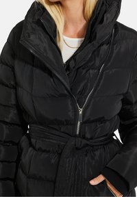 Mujer con una chaqueta acolchada negra tipo puffer, de cuello alto, cremallera diagonal, cintura ceñida con cinturón y una mano en el bolsillo con cremallera.