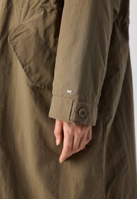 Tessuto di un cappotto trench khaki con una texture liscia, dotato di tasca a toppa, polsino con bottone e una piccola etichetta con logo sulla manica.