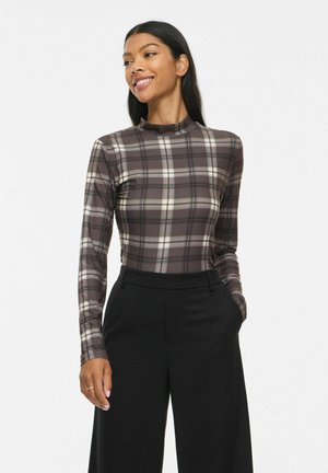 Femme portant un haut à manches longues ajusté à carreaux et un pantalon noir taille haute, souriante et regardant légèrement sur le côté, les mains dans les poches.