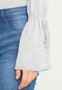 Pull gris clair avec des manches évasées, présentant des détails froncés autour des poignets. Associé à un jean en denim bleu.