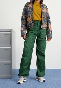 Polstret jakke i blå og brun camo mønster, sennepsfarvet t-shirt, grøn cargobukser og grå sneakers med lilla accenter.