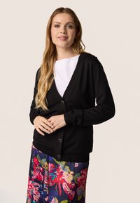 Zwart vest met een V-hals, voorzien van knoopsluitingen en geribde manchetten. Gedragen over een witte top, gecombineerd met een kleurrijke bloemenrok.