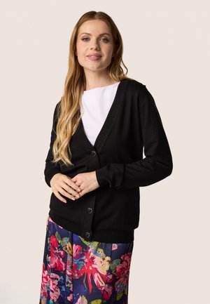 Zwart vest met een V-hals, voorzien van knoopsluitingen en geribde manchetten. Gedragen over een witte top, gecombineerd met een kleurrijke bloemenrok.