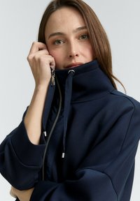 Mujer con cabello castaño claro que lleva una chaqueta azul marino con cremallera, sosteniendo la cremallera del collar cerca de su rostro contra un fondo claro.