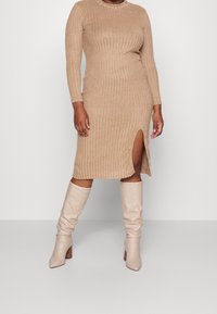 Geribbeld gebreid jurkje in lichtbeige, met lange mouwen en een zijsplit. Gecombineerd met beige, slouchy laarzen met een hoge hak tot aan de knie.