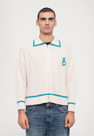 Light beige zip-up sweater with a textured weave. Features a teal collar and lower band, plus a stitched blue letter "B" on the left chest.
