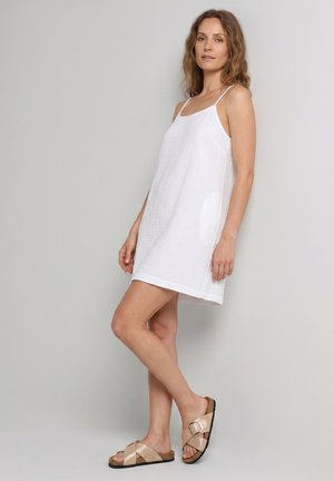 Frau in weißem ärmellosem Kleid und beigen Sandalen mit Schnalle vor hellem grauem Hintergrund.