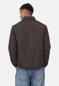 Veste isolée marron avec un col et une texture lisse, dotée d'un empiècement au dos et de poignets élastiques, associée à un jean en denim bleu clair.