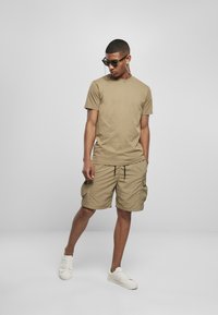 Tan bomulls t-skjorte og cargo-shorts, begge i en matchende nyanse, med justerbar liv. Modellen har på seg hvite joggesko og solbriller med kjede.