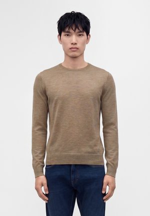 Jeune homme aux cheveux courts noirs portant un pull marron clair à manches longues et un jean bleu foncé sur un fond blanc uni.