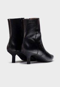 Botas de tobillo de cuero negro con una textura suave, puntera afilada y tacón delgado. Cuentan con un cierre de cremallera en la parte trasera y una marca sutil.