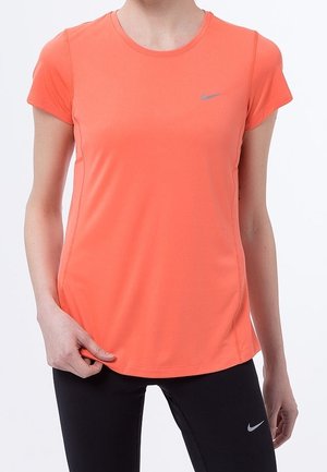 Vrouw die een feloranje sportshirt met korte mouwen en zwarte legging draagt, beide met een klein Nike-logo, staand tegen een effen achtergrond.