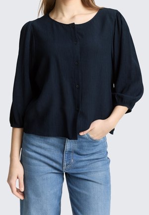 Femme portant un chemisier noir à manches longues boutonné avec des manches bouffantes et un jean bleu clair, une main dans la poche, regardant vers l'avant.
