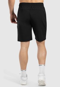 Zwarte sportshorts met een elastische tailleband, gladde stof en een losse pasvorm, getoond vanuit de achterkant. Witte sneakers en crew-sokken zijn zichtbaar.