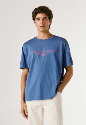 Giovane con capelli ricci che indossa una t-shirt blu Pepe Jeans London e pantaloni bianchi, in piedi contro uno sfondo chiaro e uniforme.