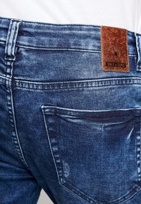 Blue denim jeans with a faded finish. Features a brown leather tag at the back waistband. Includes two back pockets and belt loops.