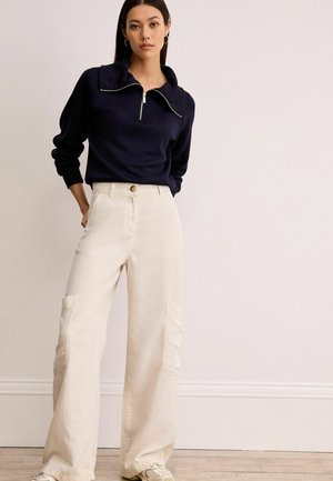 Pulover navy cu fermoar și guler înalt, cu manșete ribbate, asortat cu pantaloni largi crem, cu buzunare tip cargo și închidere prin buton frontal.