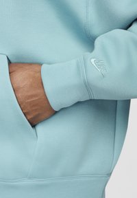 Šviesiai mėlynas Nike džemperis su minkšta tekstūra, turintis kengūros kišenę ir išsiuvinėtą logotipą ant rankogalių. Artimas vaizdas į detales.