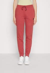 Kvinna som bär röda Nike-träningsbyxor med dragsko i midjan och vita sneakers, stående mot en enkel ljus bakgrund.