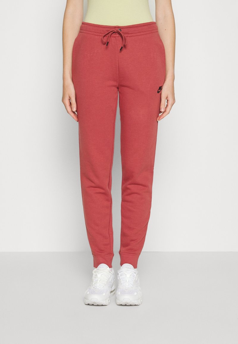 Kvinna som bär röda Nike-träningsbyxor med dragsko i midjan och vita sneakers, stående mot en enkel ljus bakgrund.