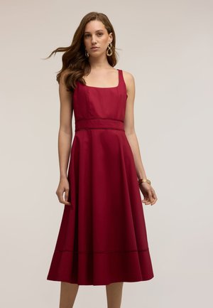 Vrouw met lang haar die een mouwloze bordeaux midi-jurk draagt, gouden creooloorbellen en gouden armbanden tegen een neutrale achtergrond.