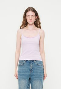 Jeune femme aux longs cheveux ondulés portant un caraco violet clair orné de dentelle et un jean bleu, debout devant un fond blanc uni.