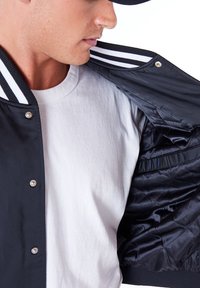 Chaqueta de varsity negra con cuello de rayas blancas, forro acolchado expuesto y botones a presión. La tela interior tiene una textura suave.