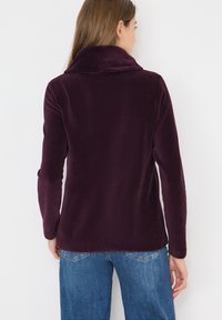 Lila Fleece-Jacke mit hohem Kragen, langen Ärmeln und weicher Textur. Der Rücken hat einen geraden Schnitt und minimale Details.