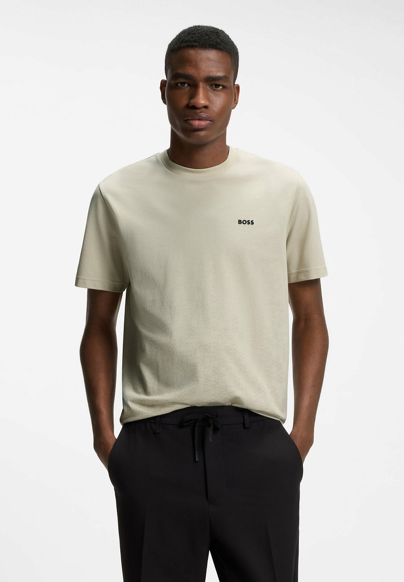 Jeune homme portant un t-shirt beige BOSS et un pantalon noir, debout les mains dans les poches devant un fond blanc uni.
