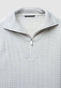 Weißer, gestrickter Pullover mit hohem Kragen und kurzem, silbernem Reißverschluss, der am Kragen teilweise geöffnet ist, mit strukturiertem Strickmuster im gesamten Pullover.
