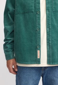 Groene corduroy overhemd met twee borstzakken, knoopsluiting aan de voorkant en ribbeltextuur. Een wit T-shirt en blauwe spijkerbroek zijn zichtbaar daaronder.