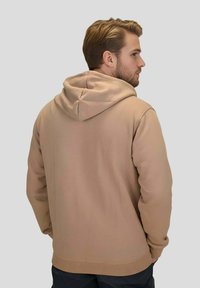 Sweat à capuche beige avec une surface texturée, doté d'une grande capuche, de poignets et d'un ourlet côtelés. Il présente une coupe ample et un design simple sans ornement.