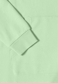 Světlé zelené rukáv mikiny s žebrovaným lemem odpočívající u velké přední kapsy typu klokánek, zobrazující texturu látky a detaily švů.