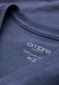 Tmavě modré bavlněné tričko s kulatým výstřihem. Etiketa uvádí "ombre bavlněná kolekce," 100% čistá bavlna, velikost M, s pokyny k údržbě.