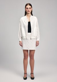 Veste bomber blanche en eyelet avec une fermeture éclair à l'avant, associée à une jupe assortie. Haut noir en dessous et chaussures noires avec des accents dorés.