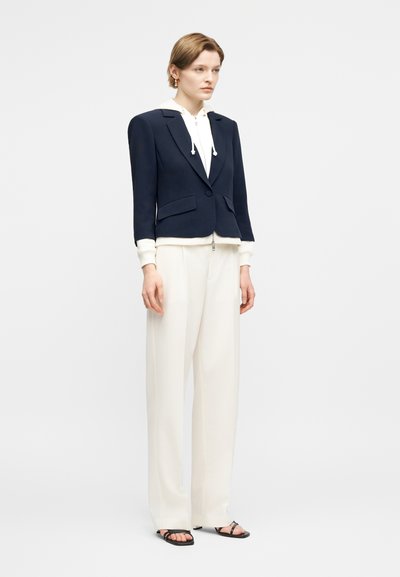 Woman wearing a navy single-button blazer over a white hoodie, white wide-leg pants, and black open-toe sandals, standing against a plain background.